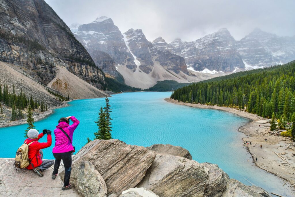 Two people dressed in warm hiking gear stand at the edge of a cliff overlooking a lake with bright blue water that is edged by green pine trees with snow-covered mountains in the background.  