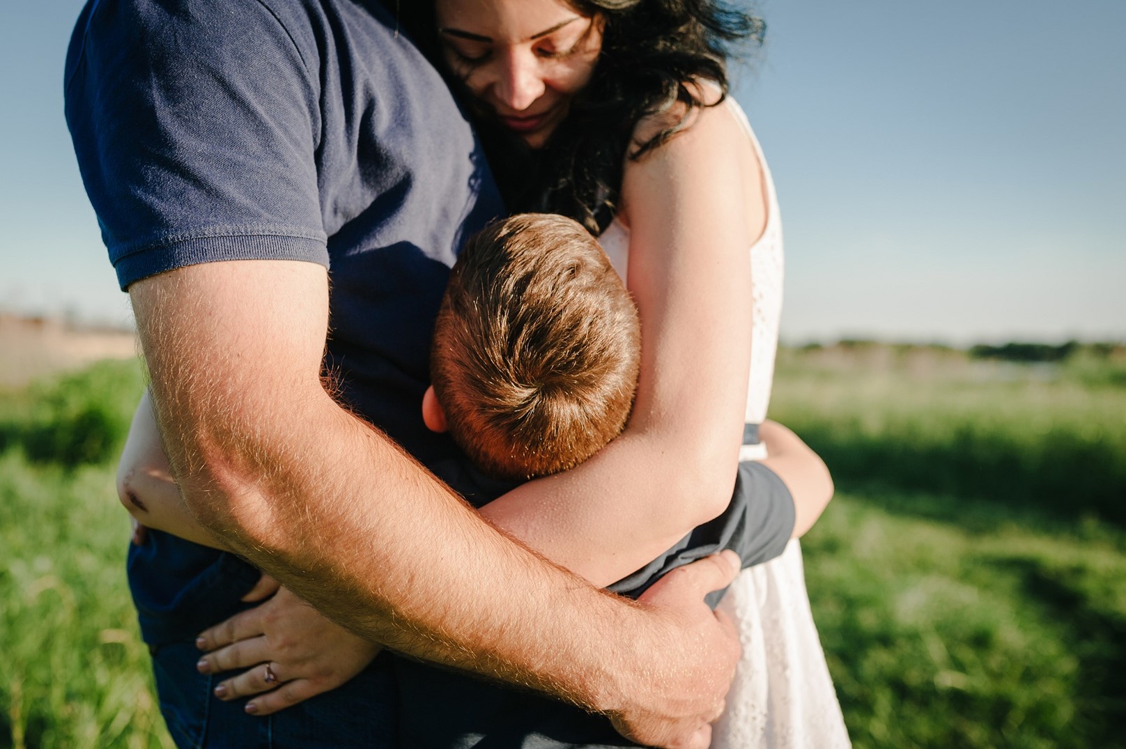 The torso of a man, a woman looking down at a young boy, and a boy looking up at the woman embrace while standing in a field 