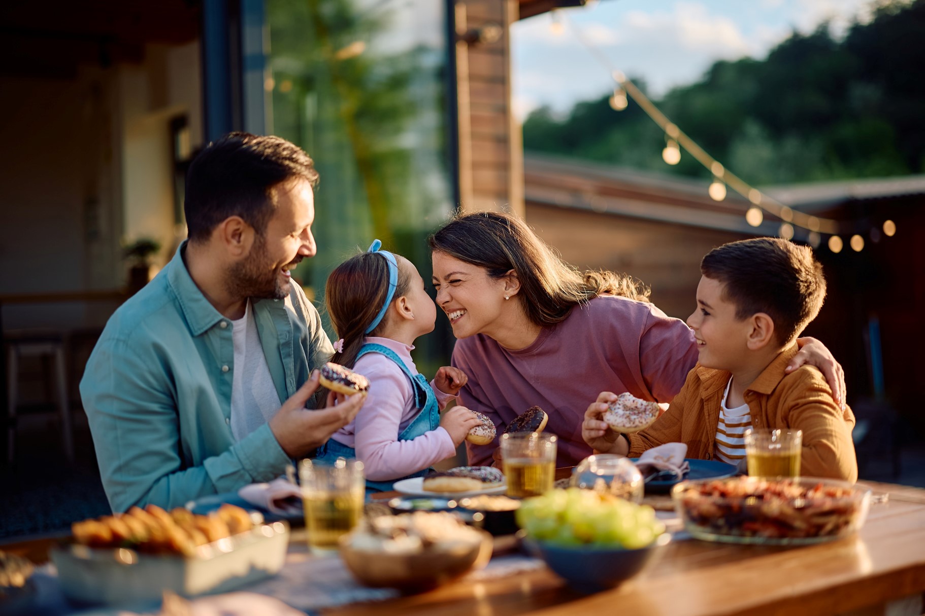 A happy dad, daughter, mom, and son sit outside at a table eating, the daughter and mom are touching their noses together. 