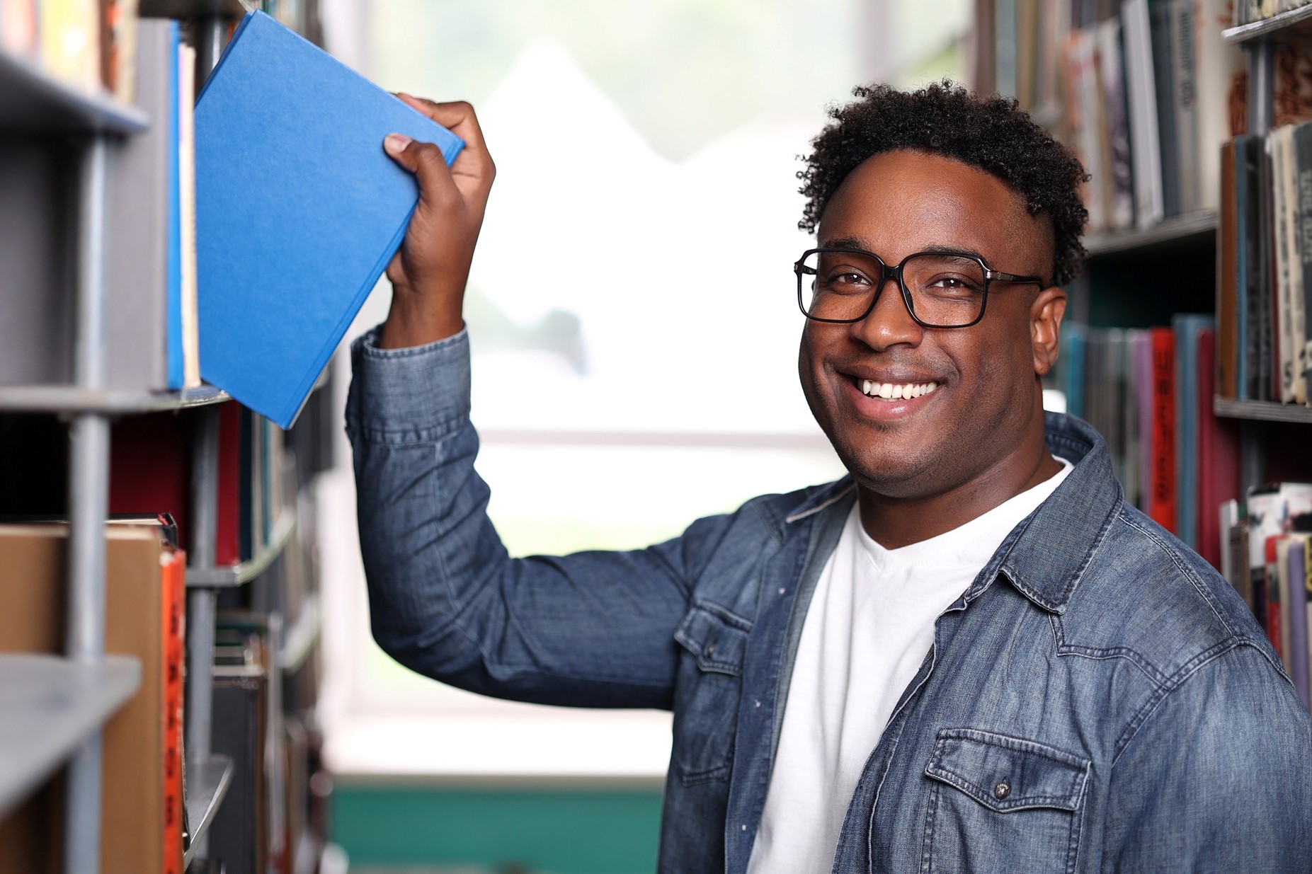 A smiling college-aged man in glasses pulls a blue book from a shelf in a library or bookstore. 