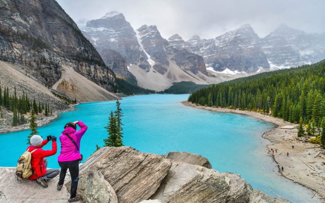 Two people dressed in warm hiking gear stand at the edge of a cliff overlooking a lake with bright blue water that is edged by green pine trees with snow-covered mountains in the background.